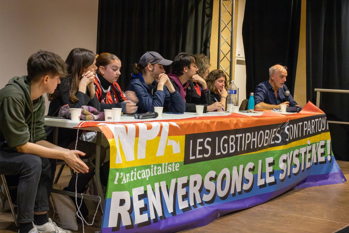 Photo de la tribune composée de 8 personnes. On peut lire sur la banderole accroché à la table "Les LGBTIphobies sont partout, renversons le systeme ! NPA"
