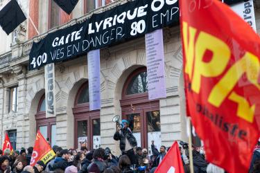 Banderole Gaité lyrique 400 vies en danger, 80 emplois menacés, un drapeau NPA sur le côté flou gaussien, un homme au mégaphone et le doigt levé au centre 