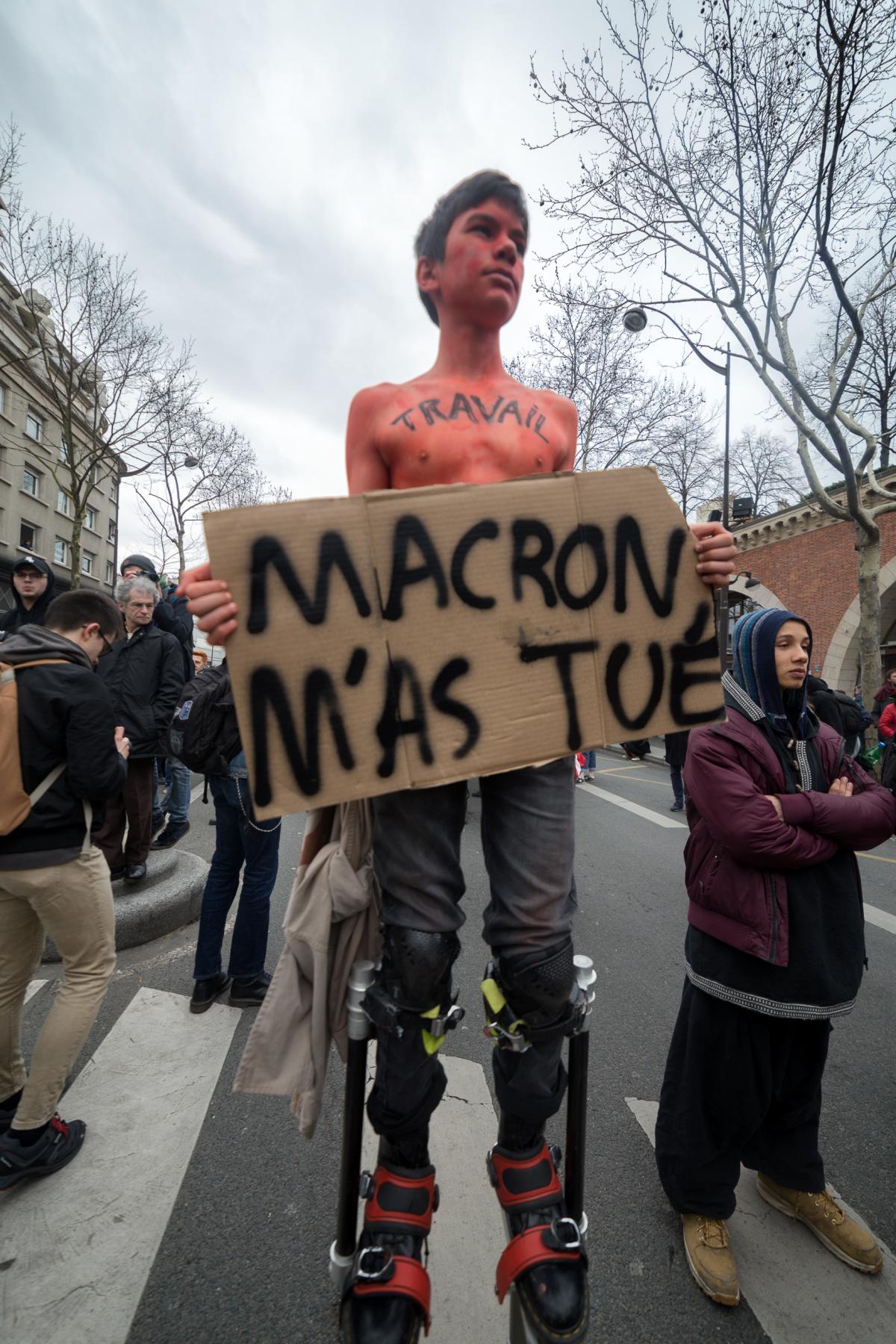 Un jeune en échasses avec une pancarte Macron M'as tué