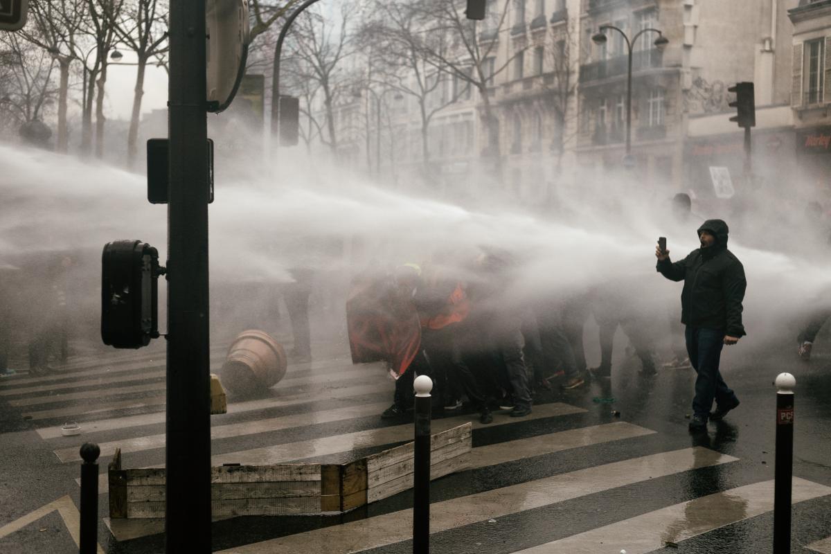 Des manifestants se protègent du canon à eau derrière une banderole, un autre filme
