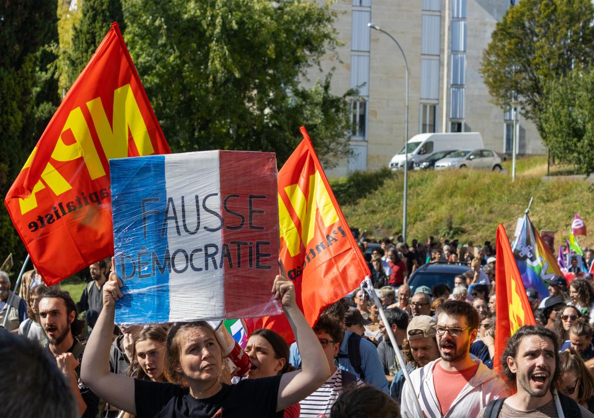 Pancarte aux couleurs du drapeau francais avec le slogan "Fausse démocratie". Des manifestants tiennent des drapeaux NPA. Manifestation contre le coup de force de Macron.